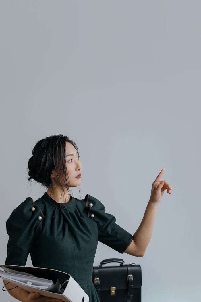 Asian businesswoman holding documents, pointing at a blank wall indoors in a minimalistic setting.