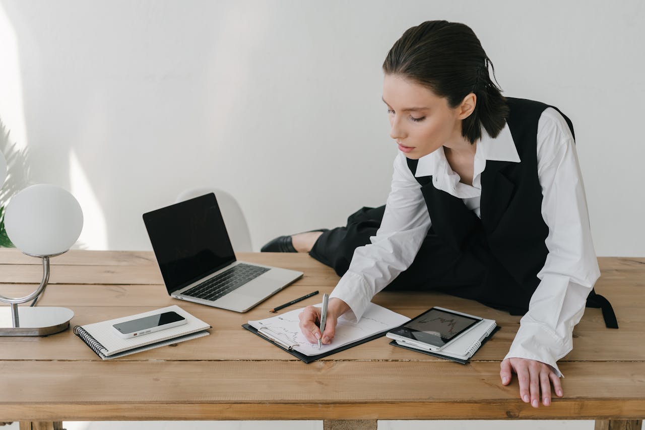 Professional woman multitasking at a desk with laptop and tablet in a modern office setting.