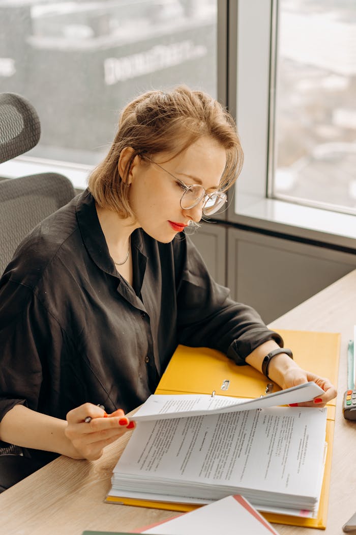 A woman in glasses sits at a desk reading documents in a modern office.